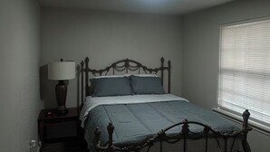 A bedroom with a bed, a nightstand, a lamp, and a window with blinds. - Home Away From Home II (Amarillo)