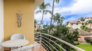 Ein Balkon mit einem weißen Korbstuhl-Tisch und -Stühlen, einem Wandbehang aus Makramee und Blick auf Palmen und Gebäude.