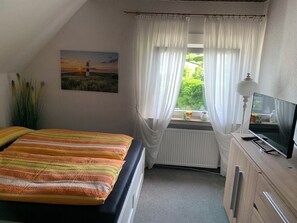 Un dormitorio con cama, ventana con cortinas, radiador, televisor y lámpara.