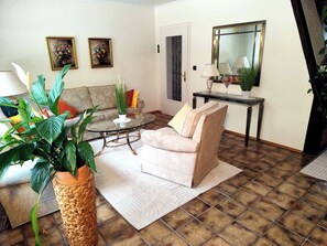 Una sala de estar con una mesa de centro de vidrio, un sofá, una planta en maceta, un cuadro y un espejo.