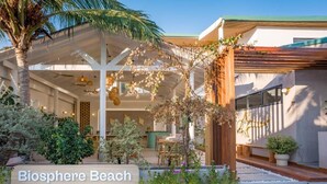 Un espacio al aire libre cubierto con una pérgola de madera, un letrero que dice 'Biosphere Beach' y diversas plantas y árboles.