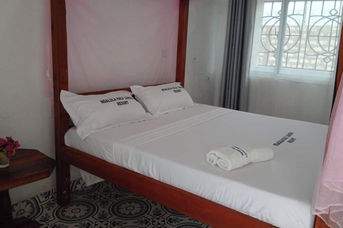 Una cama individual con sábanas blancas, dos almohadas con texto, una toalla enrollada, una mesita de noche con un florero y una ventana con celosía metálica.