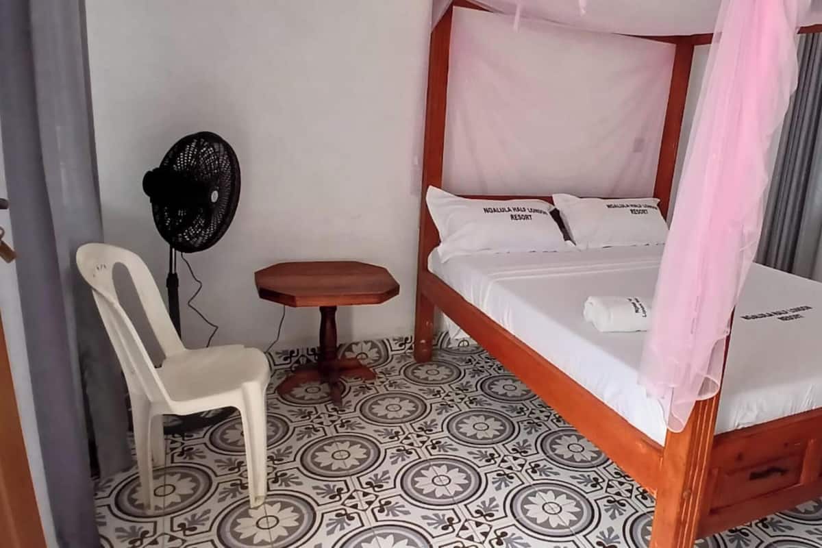 Una habitación con una cama con dosel, un taburete de madera, una silla blanca y un ventilador de pie.