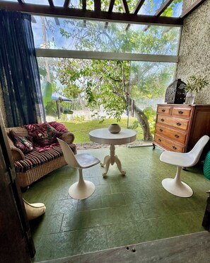 Ein Zimmer mit einer großen Glastür, die zu einem Garten führt, ein Korbsessel, ein runder Tisch mit einer Vase und eine Holzkommode.