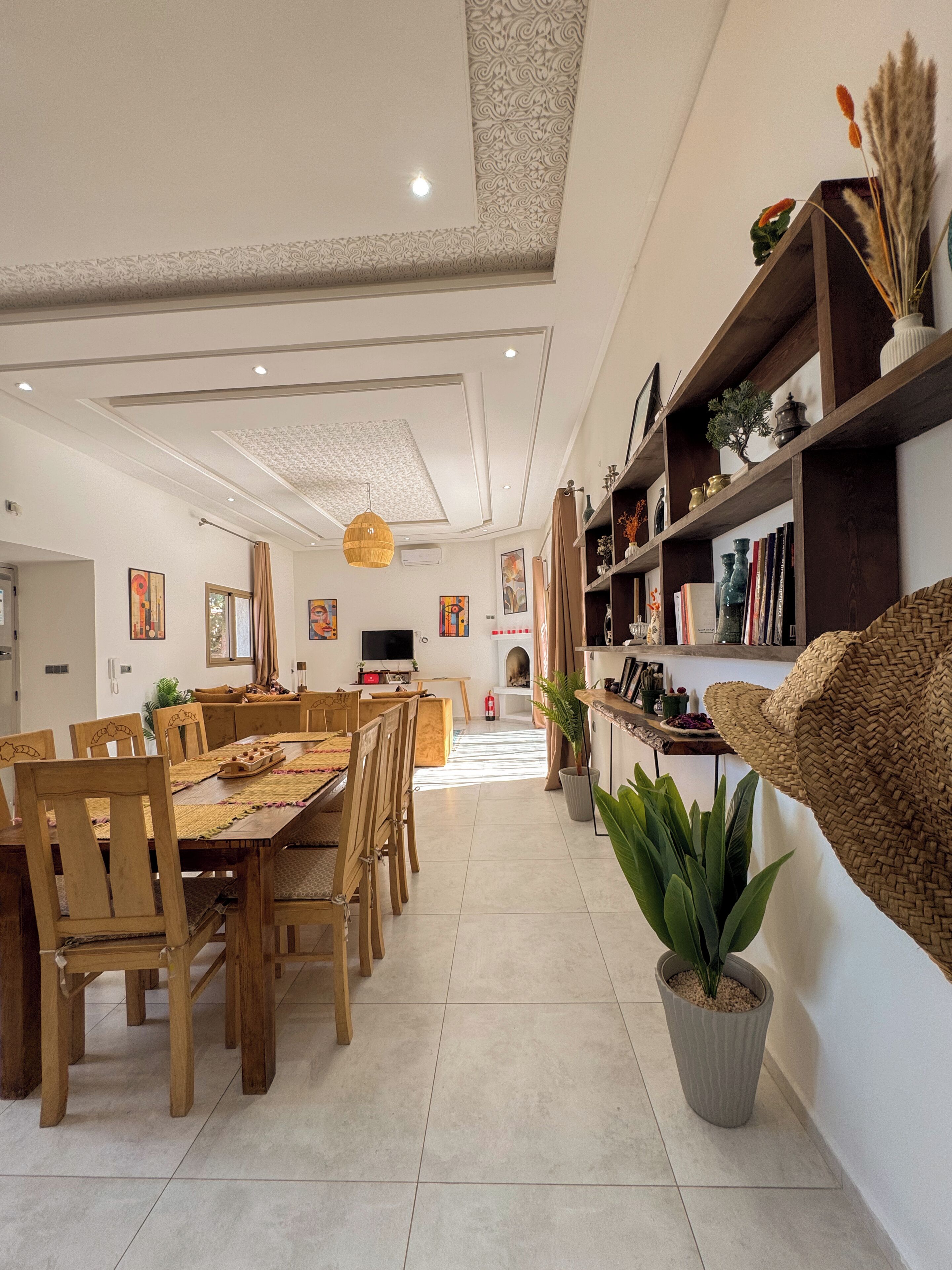 Un grand espace dédié à la salle à manger, avec une longue table en bois et des chaises, un téléviseur et une bibliothèque contenant divers objets.