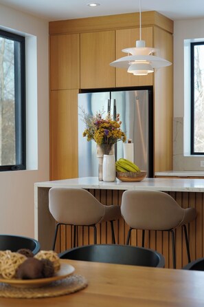 A modern kitchen with wooden cabinets, a white countertop, a stainless steel refrigerator, a vase with flowers, and a bowl of bananas. - Maison Kimby: Secluded Forest Retreat with Hot Tub (Wentworth-Nord)