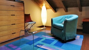 A room with a sloped ceiling, a wooden dresser, a blue armchair, a standing lamp, and a colorful rug.