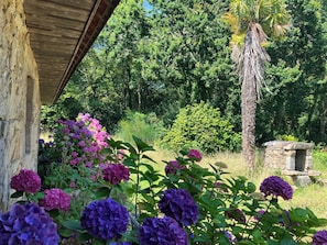 A rustic stone structure with a wooden roof, surrounded by lush greenery and vibrant hydrangea flowers. - Veritable Longére in the heart of the forest 5 minutes from the beach and all amenities (Pleumeur-Bodou)