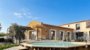 Zona de piscina amb un edifici de pedra, gandules i un parasol de palla.