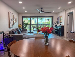 A modern living room with a dining table, sofa, piano, and a view of a balcony with palm trees. - Designer Waterfront Reno : Master Spa : Weekly Cleaning : 3-Min walk to Beach (Madeira Beach)