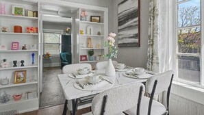 Un comedor con una mesa blanca dispuesta para cuatro personas, rodeado de estanterías blancas llenas de adornos variados.