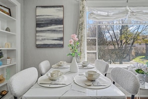 A dining area with a white table set for four, a vase with flowers, and a painting on the wall.