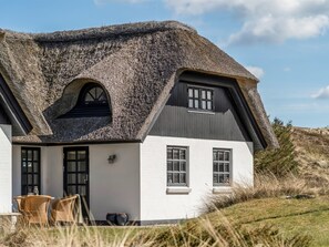 Une maison avec un toit de chaume, des murs blancs et un petit porche meublé de meubles en osier.