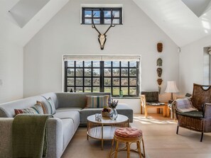 Ein großzügiger Wohnbereich mit hoher Decke, einem großen Fenster, einer Couch, einem Couchtisch, einem Korbstuhl und einem an der Wand befestigten Hirschkopf.