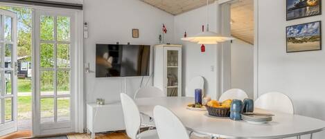 Uma sala de jantar com uma mesa redonda, cadeiras brancas, uma televisão fixada na parede e teto de madeira.