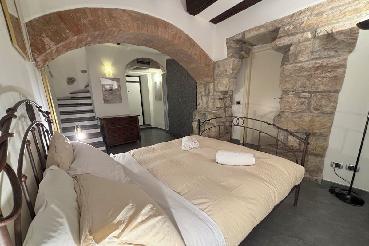 A bedroom with a stone wall, a bed with a headboard, a dresser, and a staircase leading to another room.