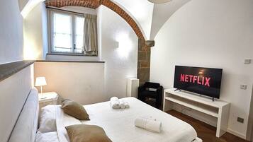 Une chambre d’hôtel avec un lit, un téléviseur affichant le logo « NETFLIX », une fenêtre avec des rideaux et un mur en pierre.