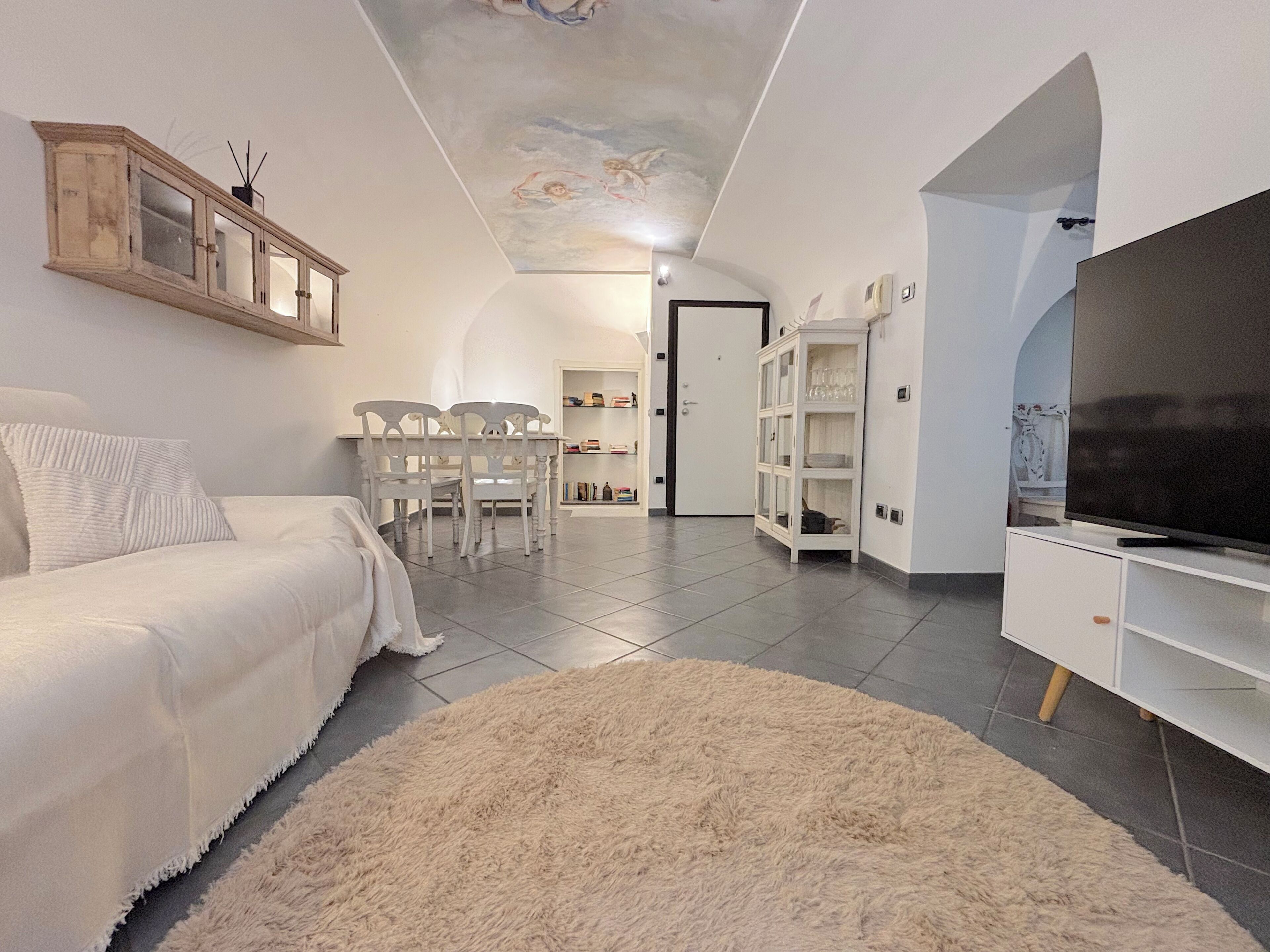 A bedroom with a bed, a TV, a white wardrobe, a dining table with chairs, and a ceiling mural.