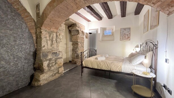 A bedroom with a stone archway, a bed with white bedding, a bedside table with a lamp, and a small round table. - Atlas Apartments - Città Alta (Bergamo)