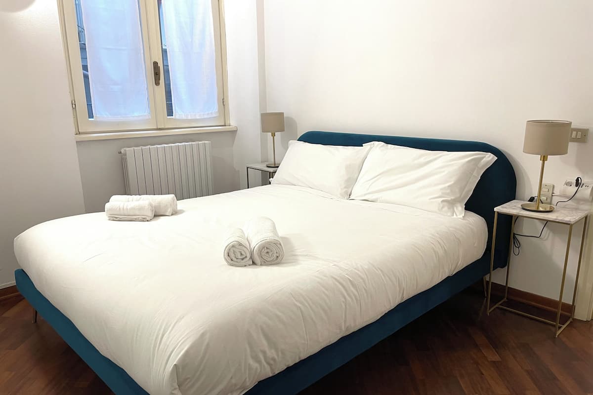 A neatly made bed with white linens and rolled towels, a blue headboard, and a bedside table with a lamp.