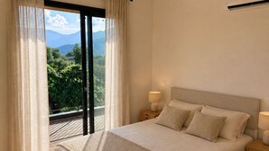 Ein Schlafzimmer mit Bett, Nachttisch, Lampe, Deckenleuchte und Blick auf Grünflächen und Berge durch ein Fenster.