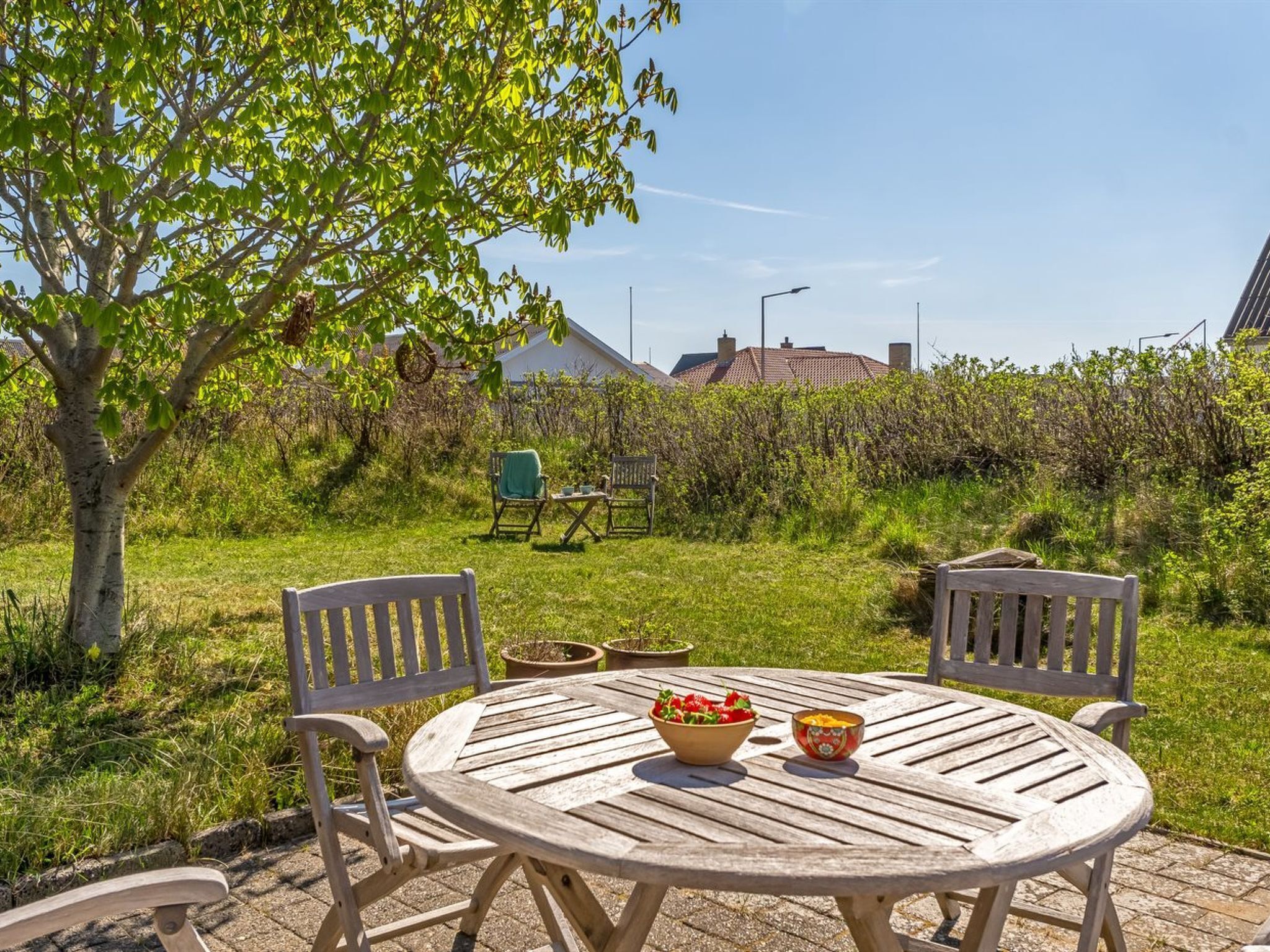 Ein runder Holztisch mit Stühlen, eine Obstschale und ein Teekessel auf einer Terrasse mit einem Baum und einem Haus im Hintergrund.