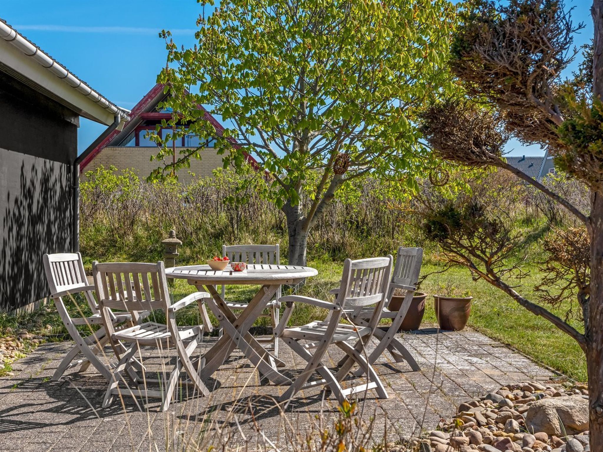 A patio with a round table and four chairs, surrounded by a garden with trees and a building in the background.