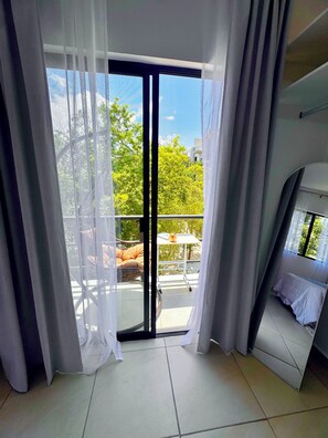 Ein Balkon mit Tisch und Stühlen, ein Bett mit weißem Kopfteil und Blick auf Bäume und Gebäude.