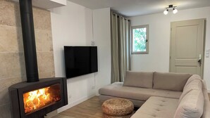 A living room with a wood-burning stove, a flat-screen TV, a beige sofa, and a woven ottoman. - Villa at the foot of Roussillon with private pool (Saint-Saturnin-lès-Apt)