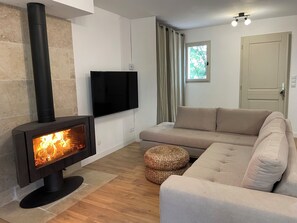 Un salon doté d’un poêle à bois, d’un téléviseur à écran plat, d’un canapé beige et d’un pouf en osier.