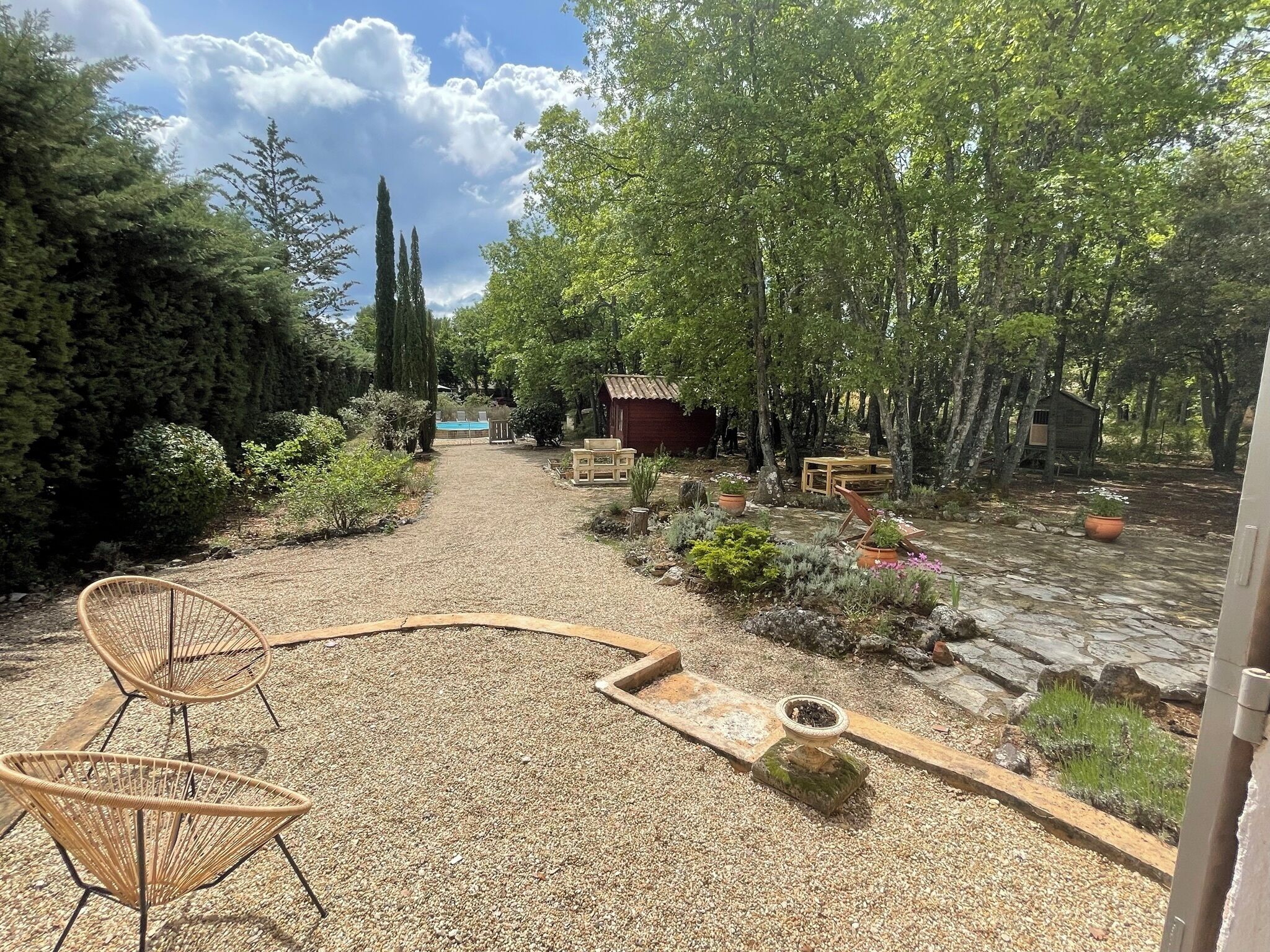 Un sendero de grava conduce a una construcción rústica de madera, rodeada de abundante vegetación y diversos maceteros.
