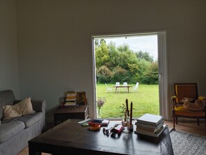 Una sala de estar con un sofá, una mesa de centro con libros y un florero, una silla y vista a un jardín a través de una puerta corrediza.