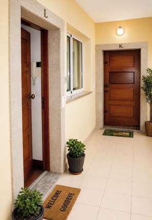 A hallway with two wooden doors, one labeled 'L' and the other 'K', a potted plant, and a welcome mat. - Pérola Apartments (Caniço)
