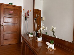 A wooden reception desk with a sign indicating an emergency exit, a mirror reflecting the room, and decorative items including a small elephant figurine and potted plants. - Pérola Apartments (Caniço)