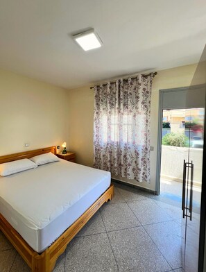 A hotel room with a bed, a nightstand, a lamp, a window with floral curtains, and a door leading outside. - Private Full Home in Agadir - Families & Groups (Agadir)