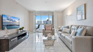 A modern living room with a sofa, a glass coffee table, a TV, and a city view through large windows. - Spectacular Water Views 2br-4 beds in Sunny Isles Beach (Sunny Isles Beach)