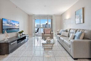 A modern living room with a sofa, a glass coffee table, a TV, and a city view through large windows. - Spectacular Water Views 2br-4 beds in Sunny Isles Beach (Sunny Isles Beach)