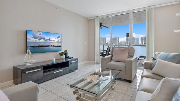 A modern living room with a glass coffee table, a sofa, a TV mounted on the wall, and a view of the cityscape through large windows. - Spectacular Water Views 2br-4 beds in Sunny Isles Beach (Sunny Isles Beach)