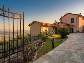 Uma casa com um caminho de pedra que leva a um portão, um jardim com diversas plantas e vista para uma cidade distante e montanhas.