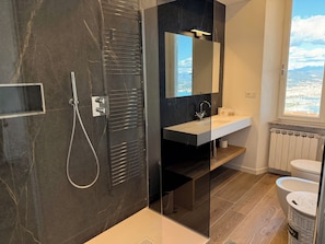 Un baño moderno con ducha de vidrio, lavamanos blanco, espejo y vistas a la ciudad a través de una ventana.