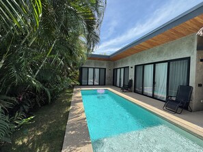 Ein moderner Poolbereich im Freien mit einem klaren, blauen Pool, umgeben von üppiger Vegetation und großen Glasschiebetüren.