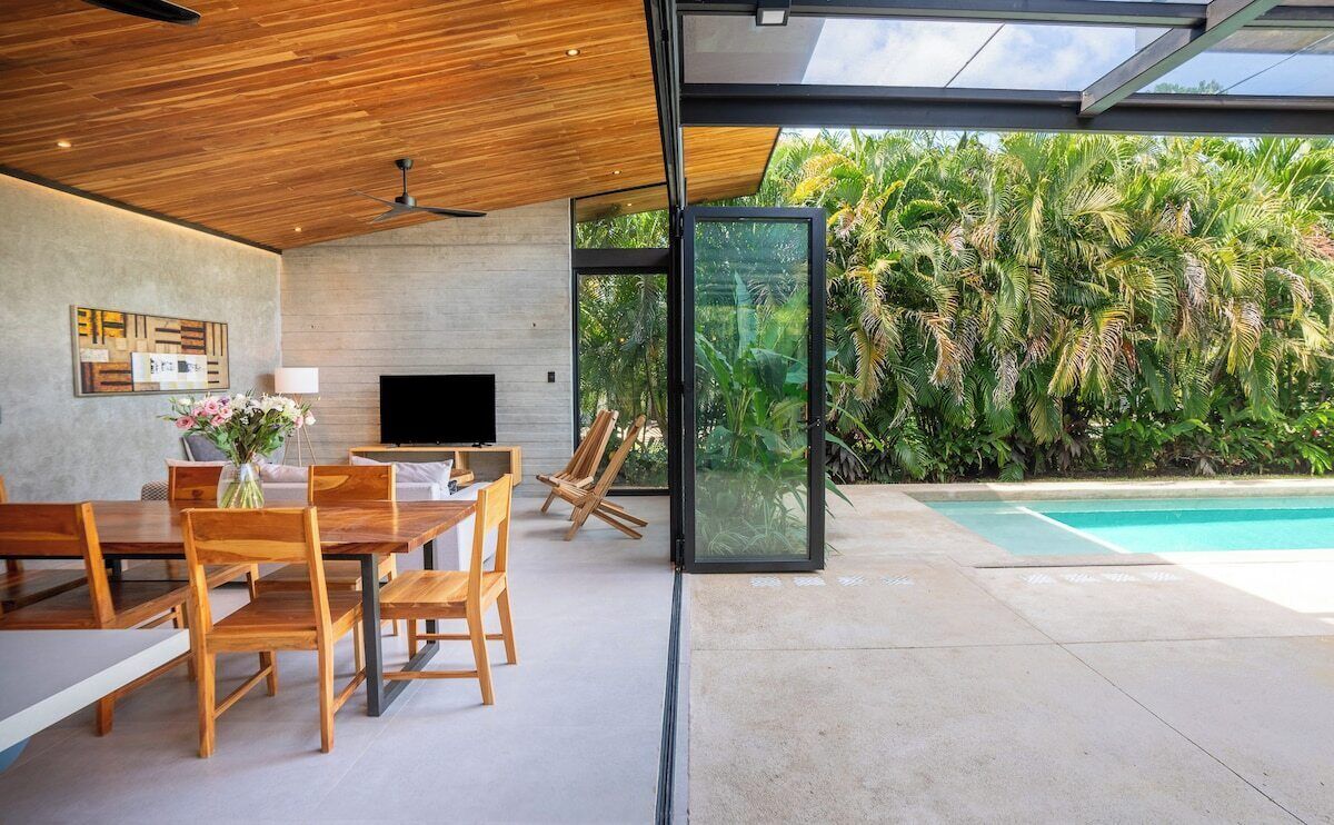 A modern interior with a dining area, a flat-screen TV, and a glass wall overlooking a pool and lush greenery.