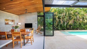 Een modern interieur met een eethoek, een flatscreen televisie en een glazen wand met uitzicht op een zwembad en weelderige begroeiing.
