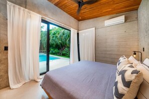 Ein Schlafzimmer mit Bett, Deckenventilator, Klimaanlage und Blick auf Pool und Grünflächen.