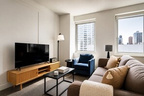 A modern living room with a flat-screen TV, a wooden entertainment unit, a blue armchair, a sofa, and a city view through large windows. - Luxury Midtown 2BR , Views, 24/7, Doorman, W/D - New York City (New York)