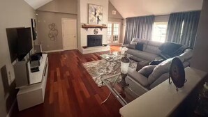 A spacious living room with a fireplace, a ceiling fan, and a large window with curtains.