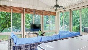 Uma sala de sol com mesa de bilhar, assentos acolchoados azuis, uma televisão e grandes janelas com vista para um lago.