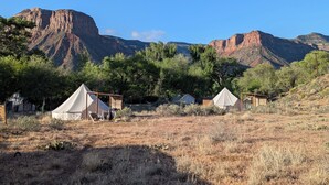 Un camping con tiendas de campaña y una cabaña de madera, rodeado de pastizal seco y árboles, con montañas al fondo.