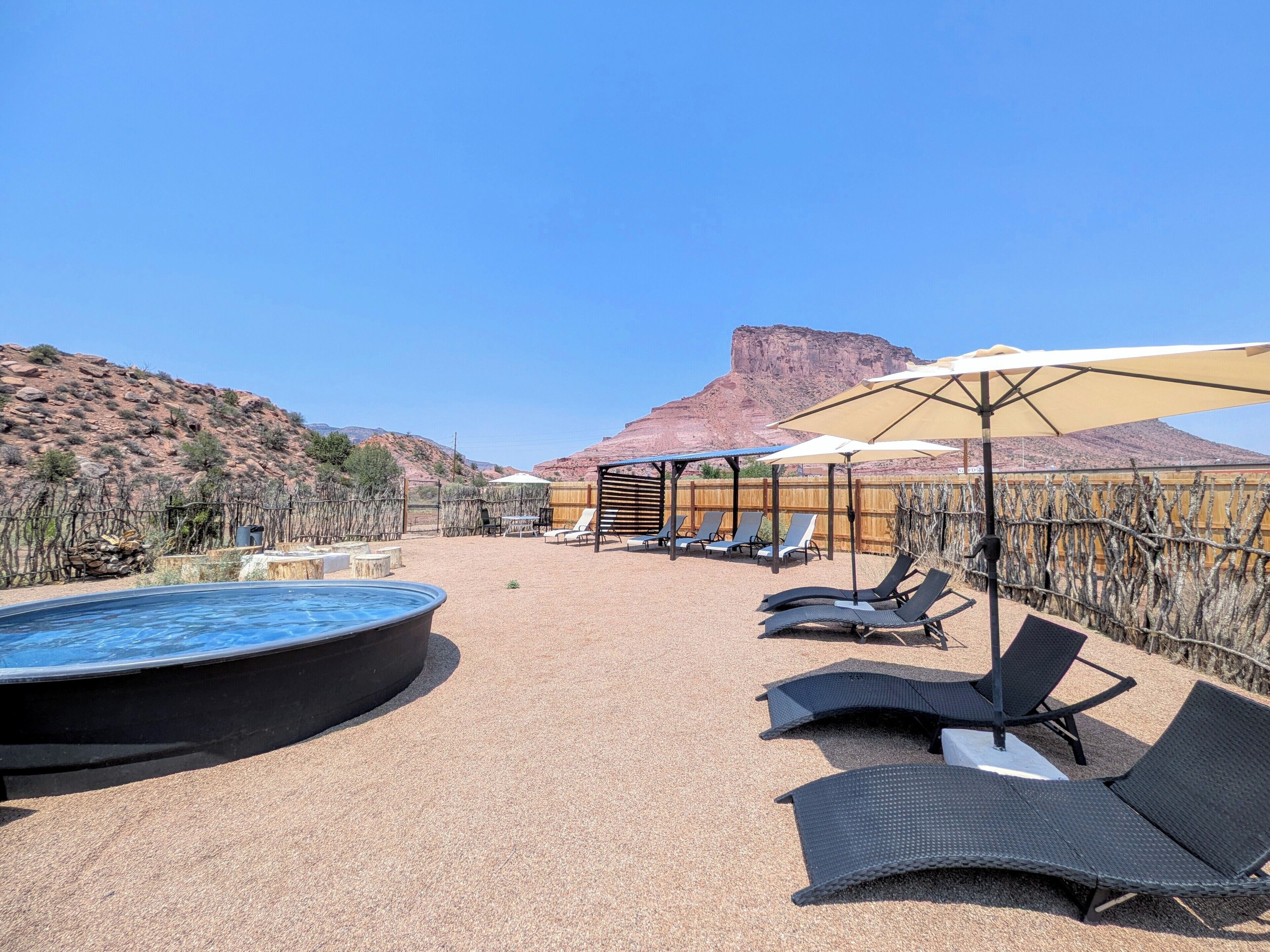 Zona de piscina con sillones de descanso y sombrillas, rodeada de vegetación desértica y una montaña al fondo.