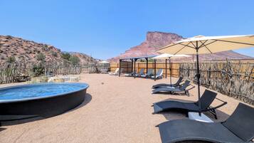 Zona de piscina con sillones de descanso y sombrillas, rodeada de vegetación desértica y una montaña al fondo.